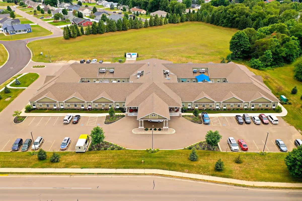 Aerial view of a single-story senior living facility named Kauhale Otsego, surrounded by parking lots, green lawns, and trees, with residential houses and wooded areas in the background.