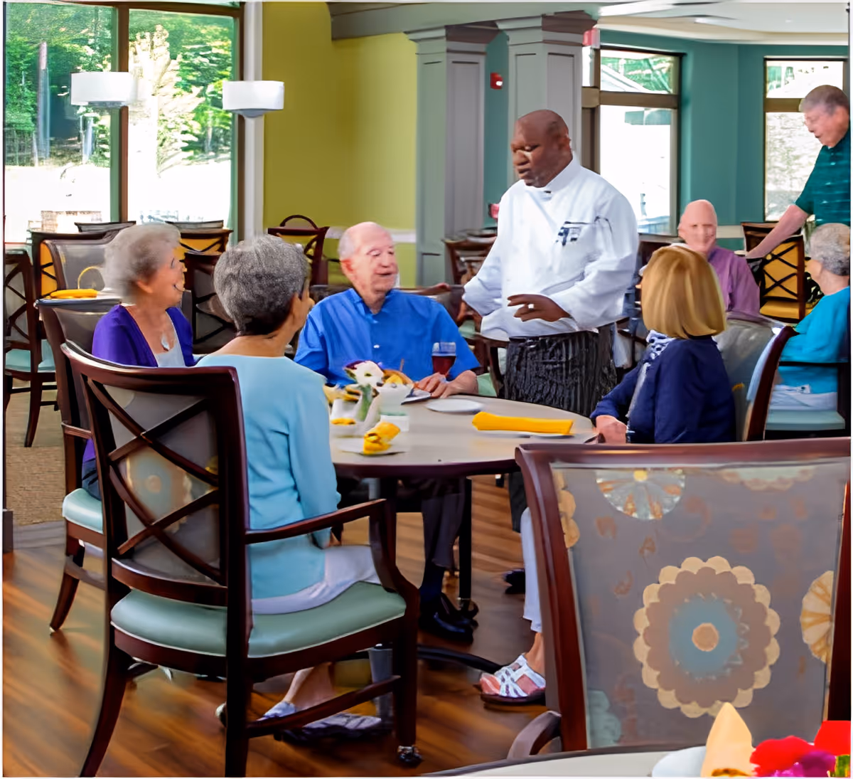 A group of elderly people seated around a dining table in a bright, spacious dining room with large windows. A chef in a white uniform is standing and interacting with the group. The room has wooden floors, cushioned chairs, and yellow napkins on the tables.