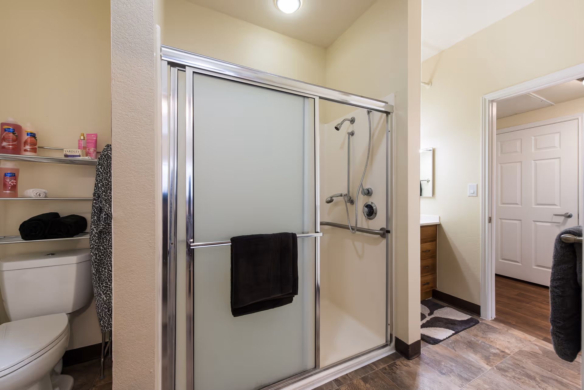 Bathroom with a frosted glass sliding shower door, a showerhead with a handheld attachment, a toilet, and metal shelves holding toiletries and towels. The bathroom has beige walls and a tiled floor, with a view into an adjacent room with wooden flooring and white doors.