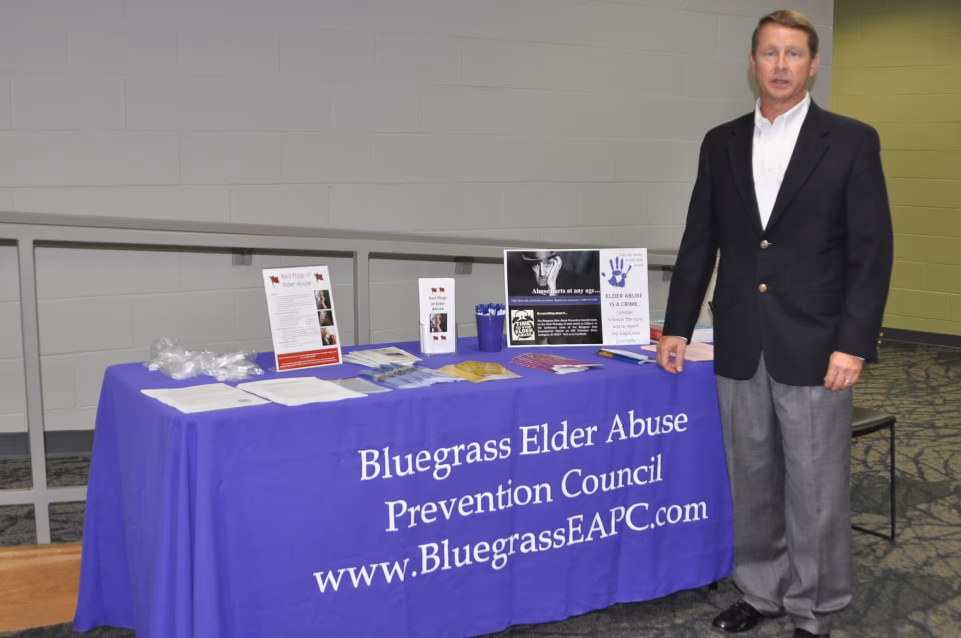 Man in a suit stands beside a purple table banner at an indoor information booth.