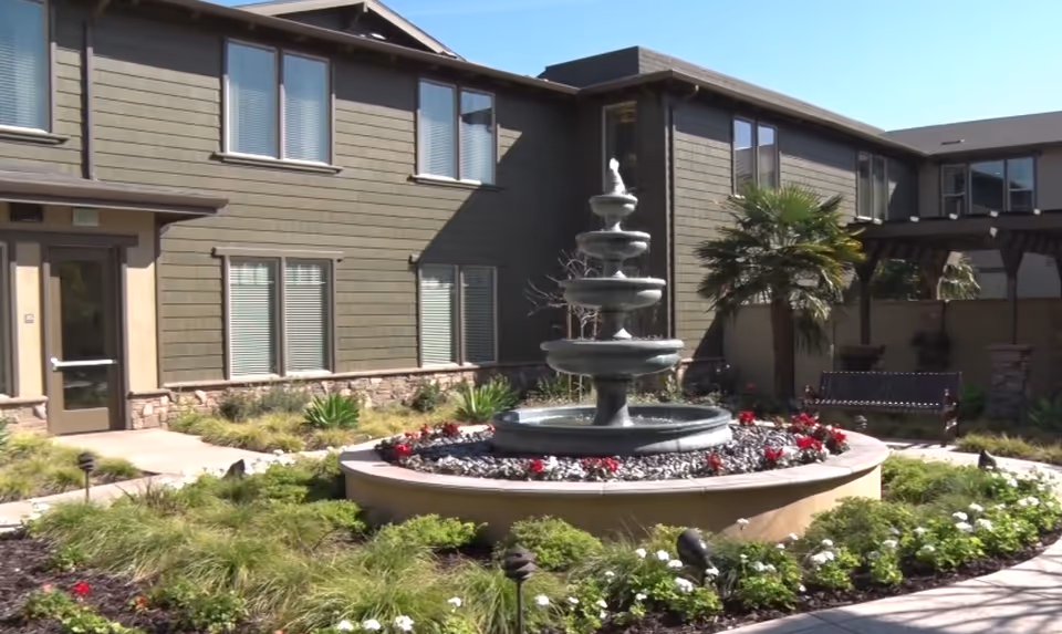 Courtyard with a multi-tiered fountain surrounded by flowerbeds and a two-story building in the background.