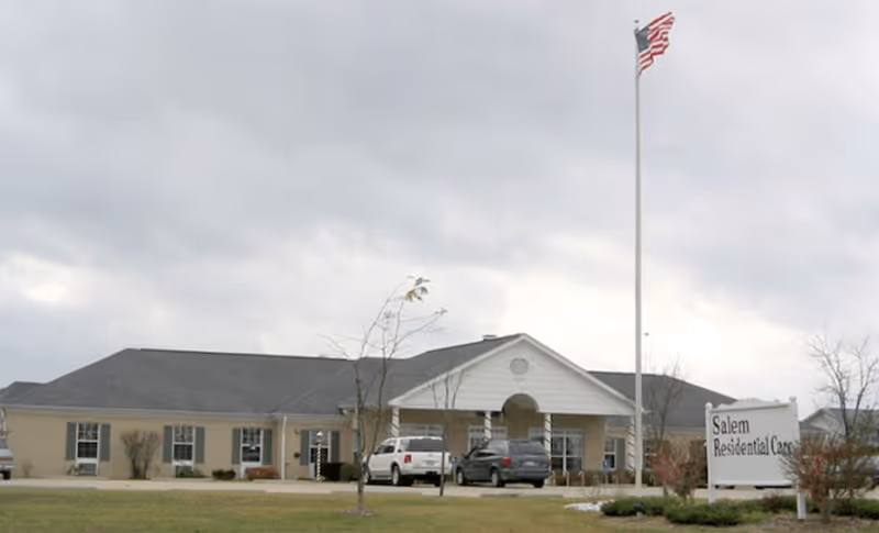 Exterior view of Salem Residential Care Facility, a single-story building with a pitched roof, several windows, and a covered entrance. There are a few cars parked in front, a flagpole with an American flag, and a sign displaying the facility's name on the lawn.