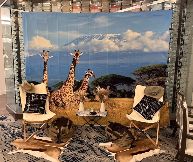 Indoor seating area with two beige folding chairs each holding a black pillow with white text and a tan hat. Between the chairs is a small table with two vases of dried flowers and a pair of binoculars. Behind the seating area is a large backdrop featuring three giraffes in a grassy savanna with trees and a mountain in the background. The floor has patterned carpet with two animal hide rugs under the chairs and bags placed on the rugs.