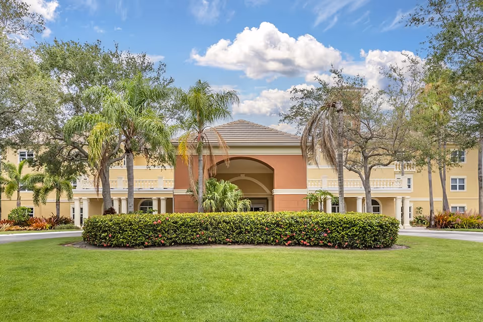 Front exterior view of Brookdale Jensen Beach facility showing a well-maintained lawn, trimmed bushes, palm trees, and a building with a covered entrance under a partly cloudy sky.