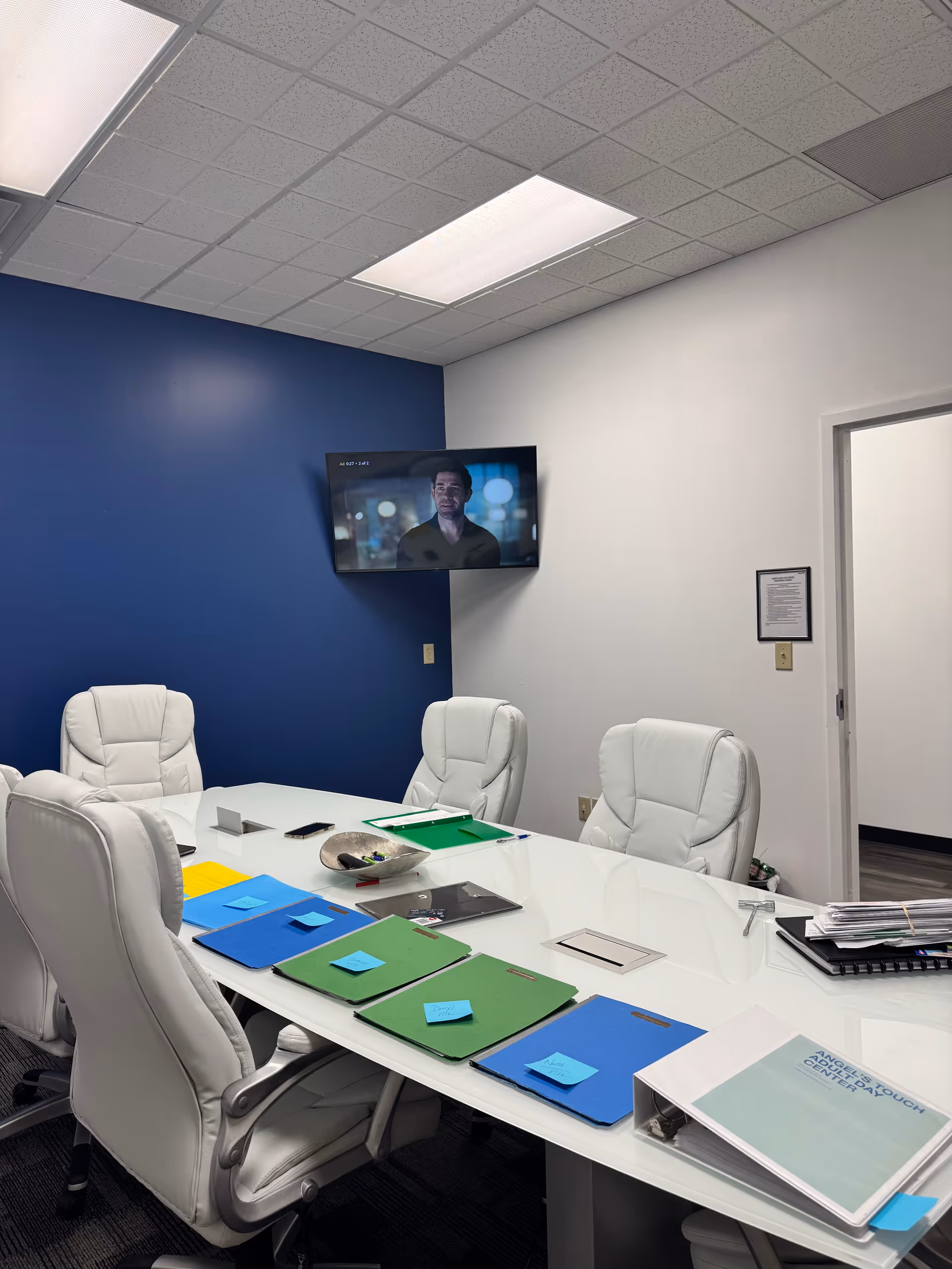 A conference room with a white table, white chairs, blue accent wall, and a mounted TV.
