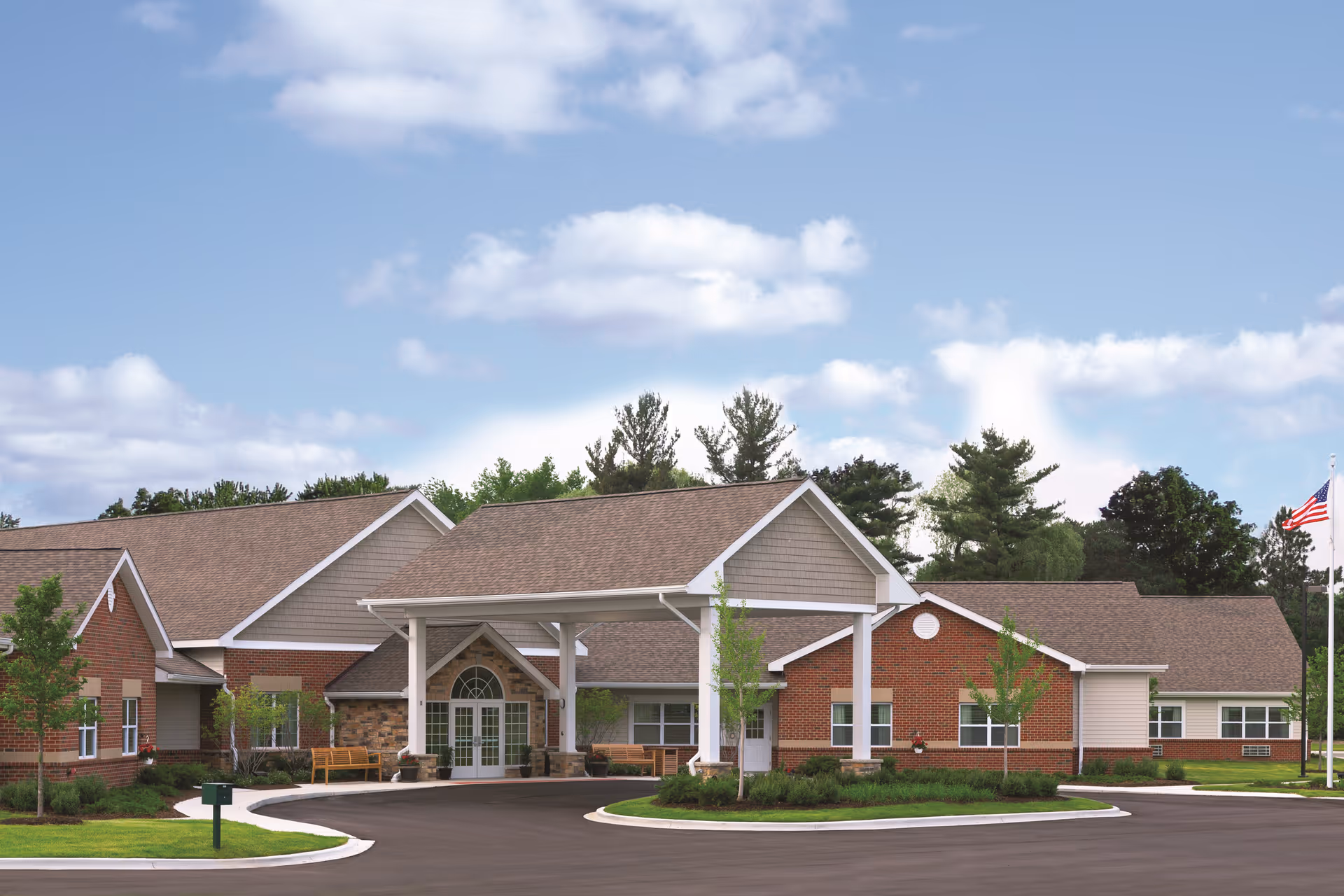 Exterior view of Brittany Manor, a single-story senior living facility with a covered entrance, brick and siding facade, surrounded by landscaped greenery and trees under a partly cloudy sky.