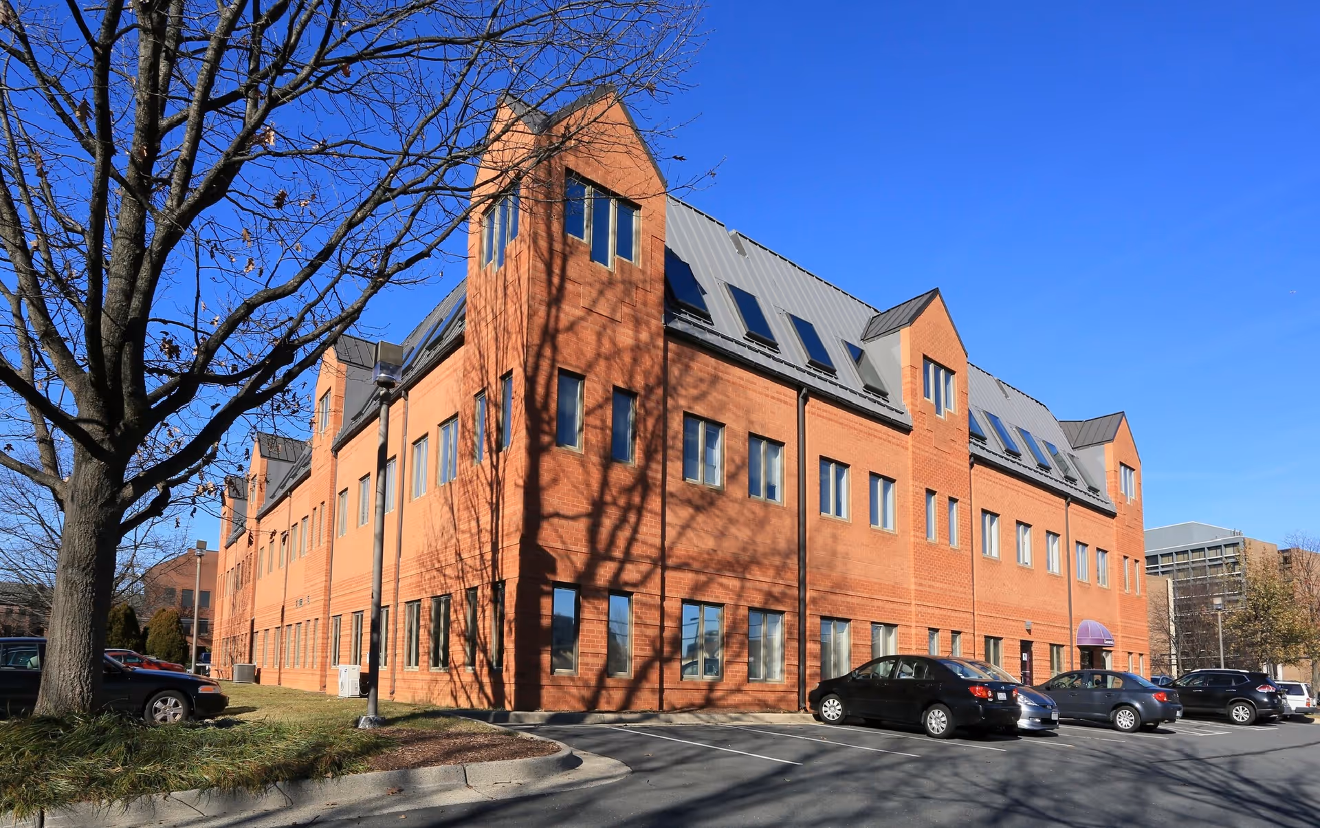 Brick senior living facility exterior with parked cars and a leafless tree against a blue sky.
