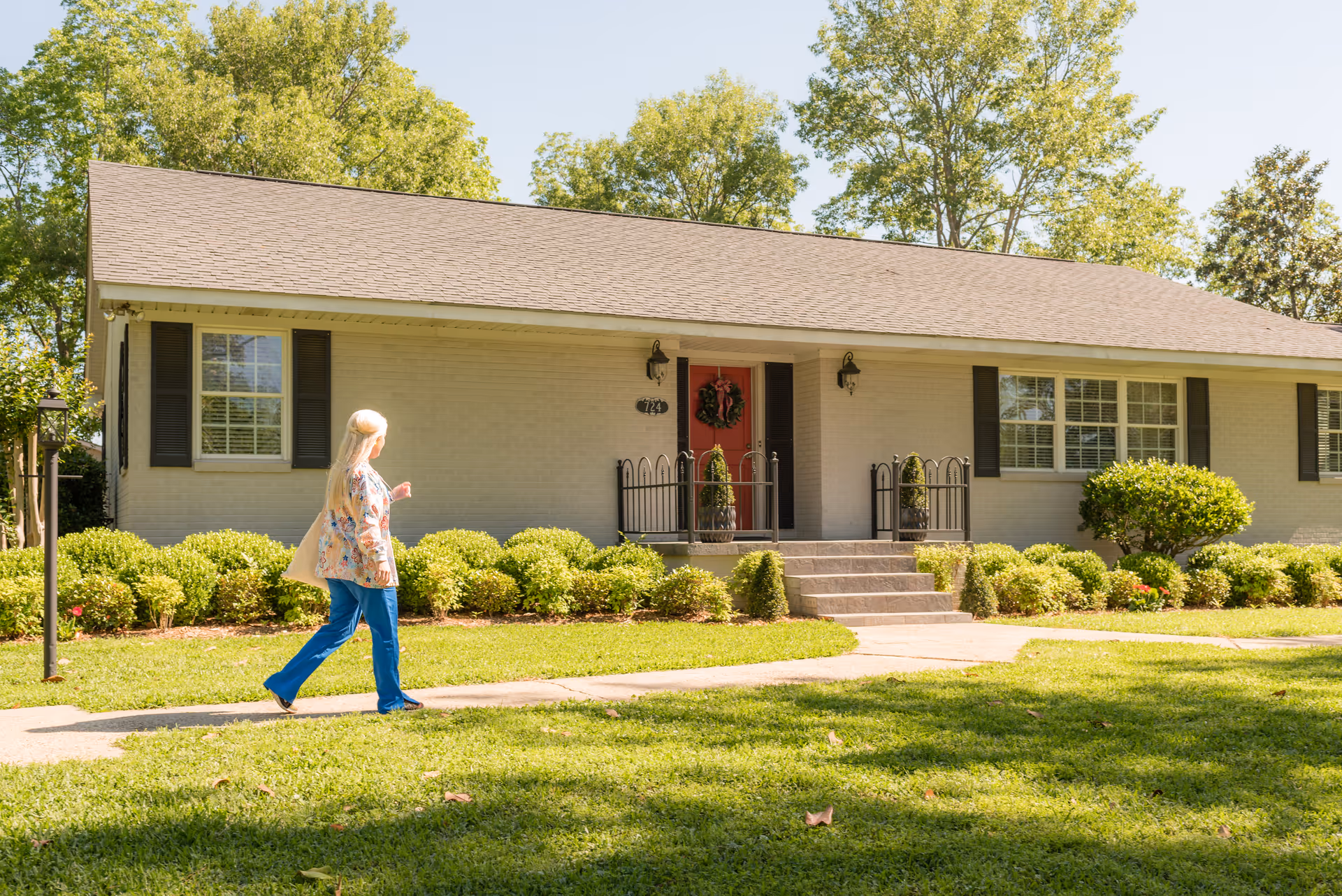 Senior living facility exterior with front entrance, shrubs, and an elderly woman walking.