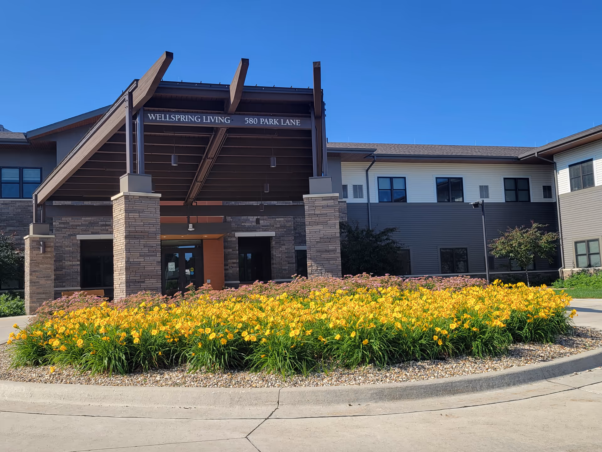 Exterior view of a senior living facility entrance with stone columns and yellow flowers