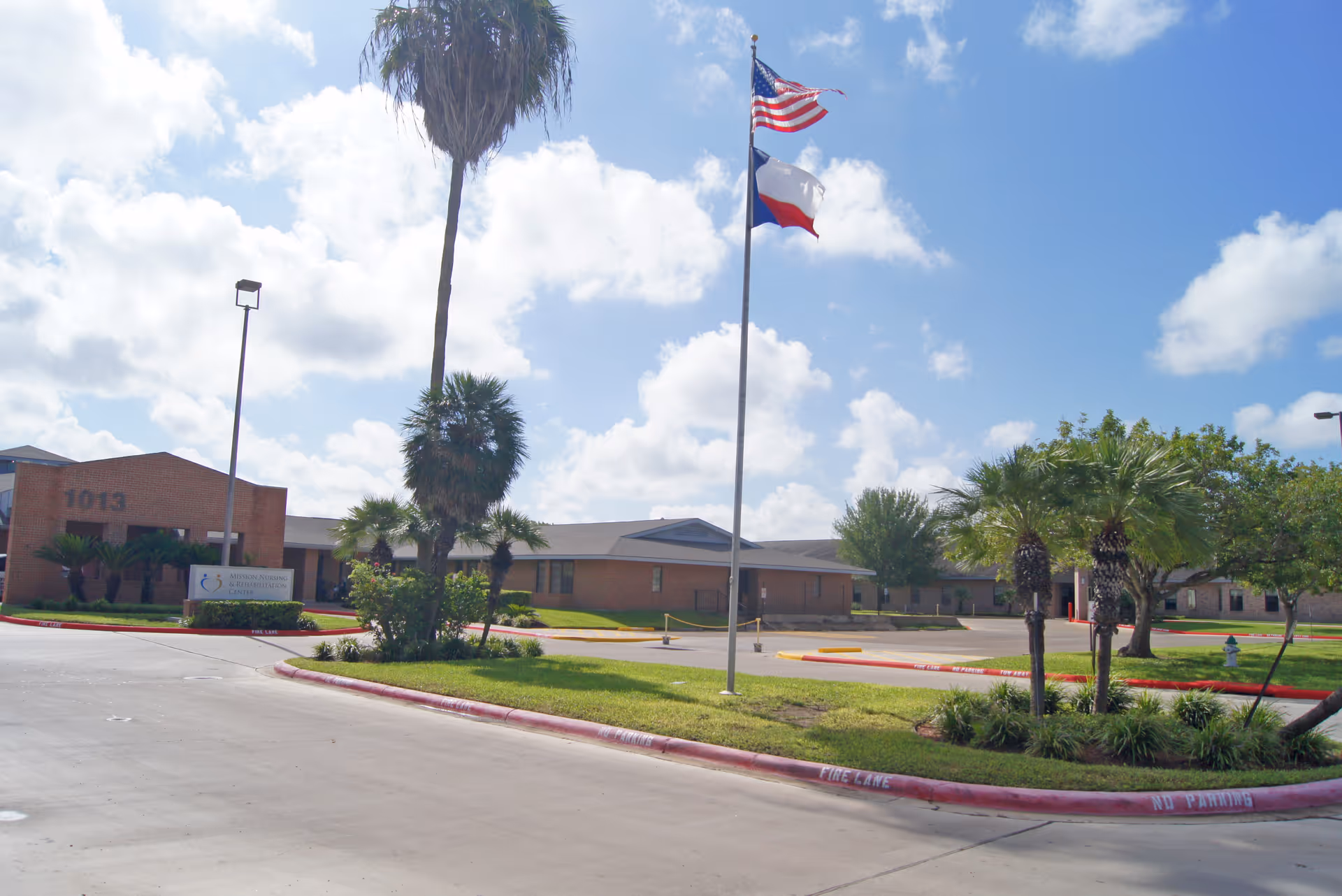 Front exterior of Mission Nursing and Rehabilitation Center with a low brick building, flagpoles flying the U.S. and Texas flags, palm trees, and a curved driveway.