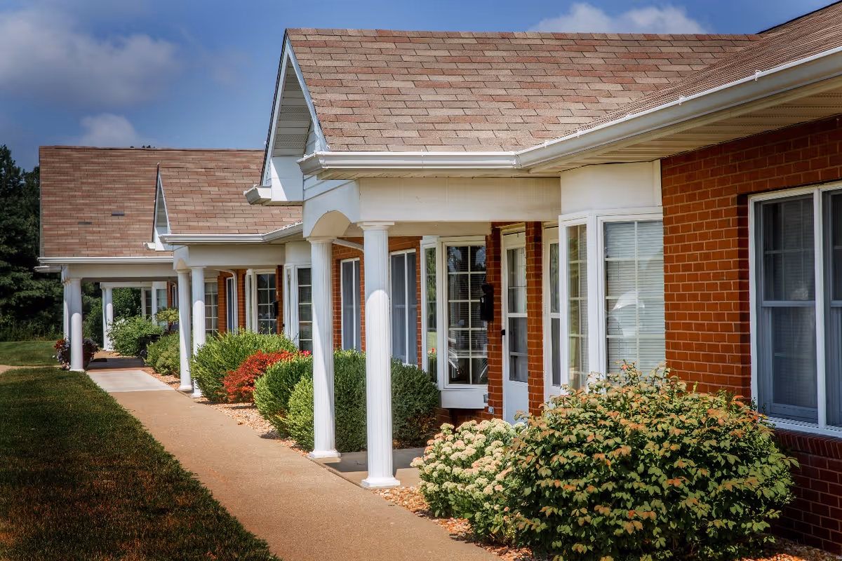 Front exterior of a single-story brick residential building with white columns, a covered walkway, and landscaped shrubs.