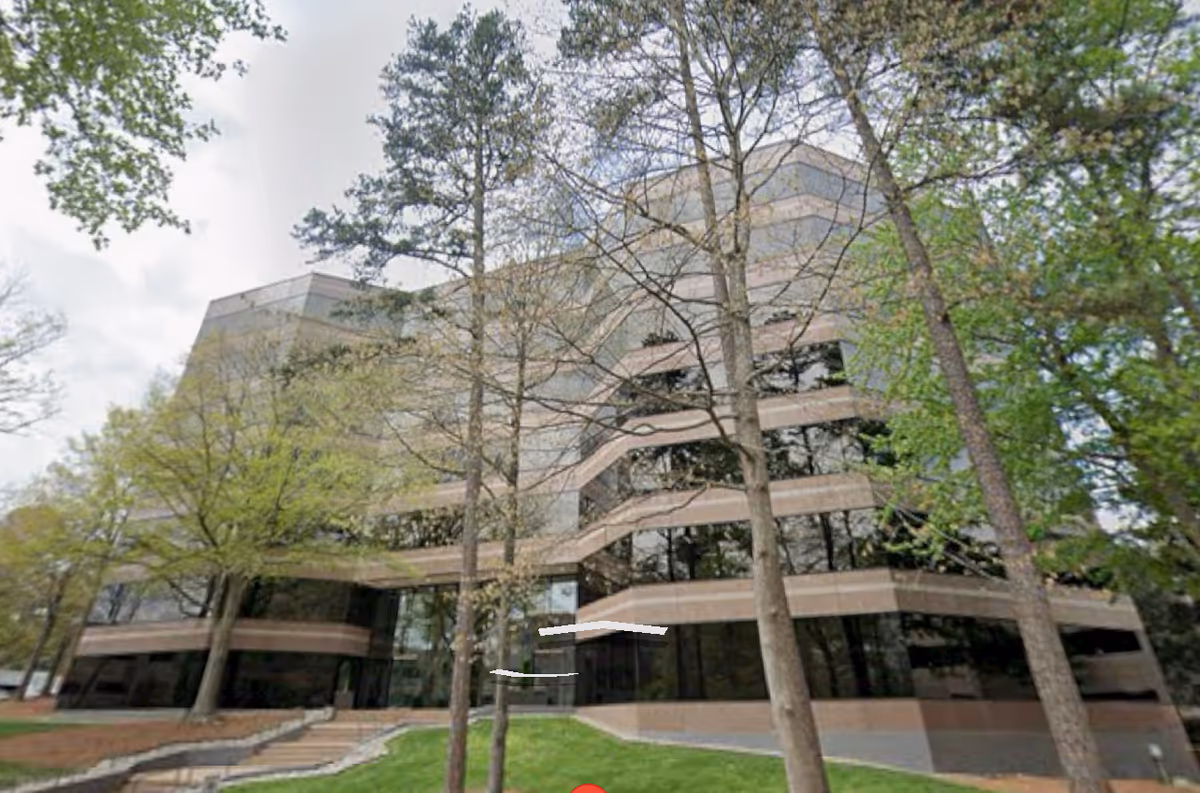 Multi-story building with glass facade, surrounded by trees, entrance stairs visible.