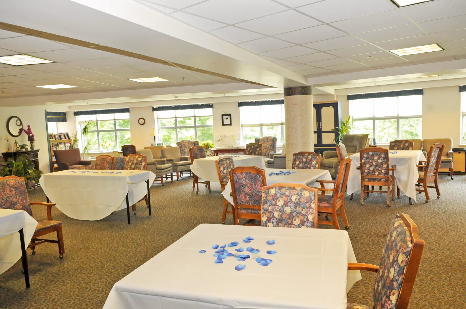 A spacious, well-lit common area with multiple tables covered in white tablecloths and decorated with scattered blue petals. The room has floral upholstered chairs, large windows letting in natural light, and various armchairs and plants around the perimeter.