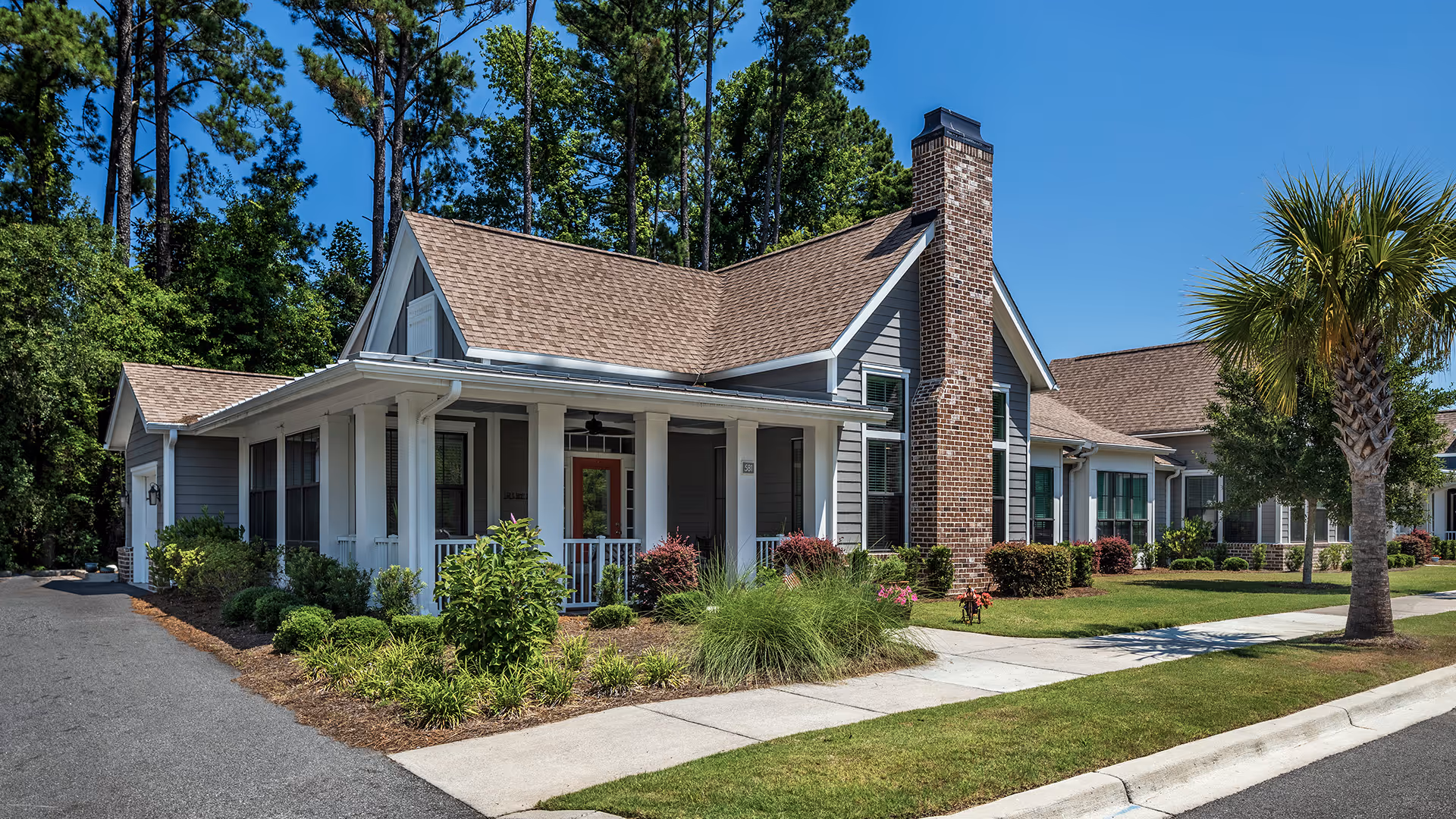 Exterior view of a single-story residential building with a gabled roof, brick chimney, and a covered front porch surrounded by landscaped greenery and a palm tree, under a clear blue sky.