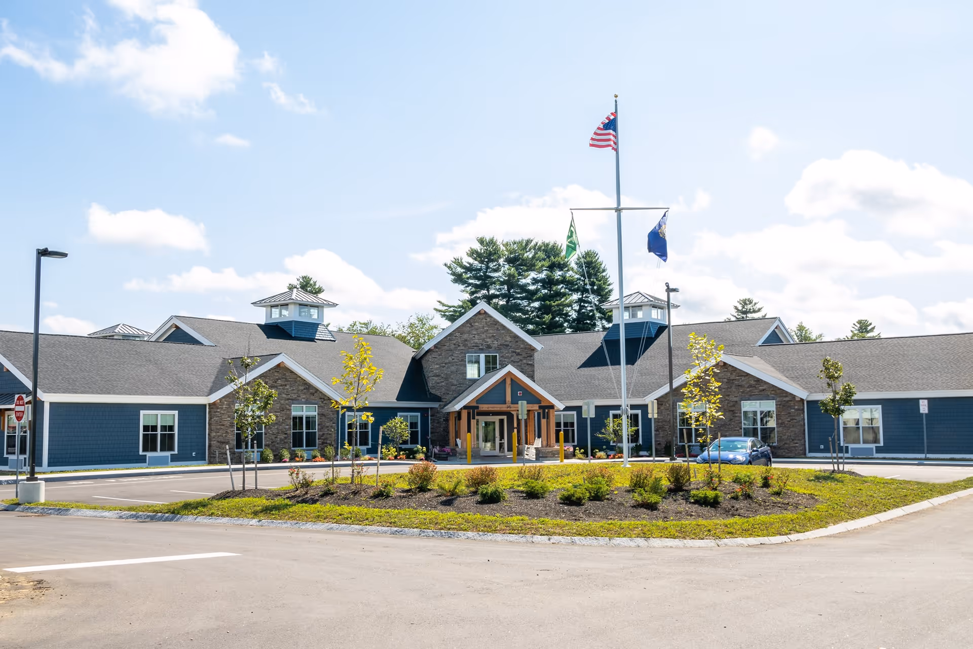 Front view of a blue senior living facility with landscaping, flagpoles, and a central entrance.