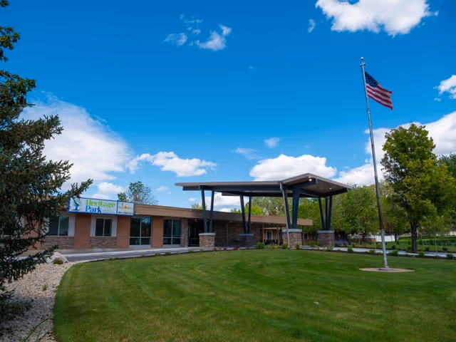 Front exterior of Heritage Park Healthcare and Rehabilitation showing a covered entrance, American flag, and manicured lawn under a blue sky.