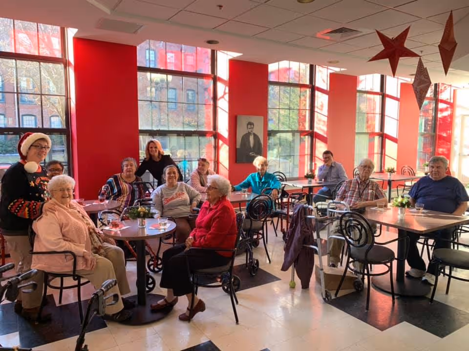 Elderly residents gathered at tables in a bright dining/activity room with large red-trimmed windows.