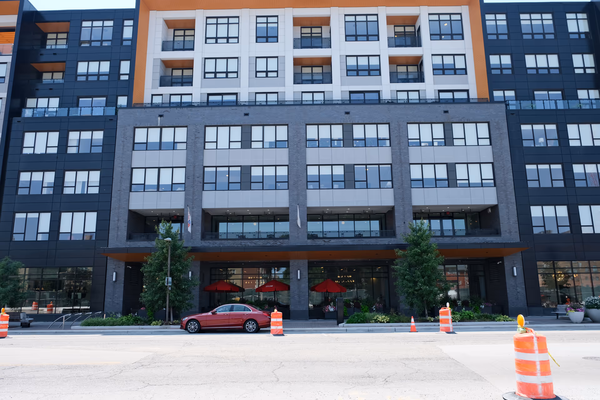 Front exterior view of a modern multi-story building with large windows and balconies. There are three red umbrellas visible on the ground floor patio area. A red car is parked on the street in front of the building, which is bordered by orange construction barrels and cones. Trees and plants are also visible near the entrance.
