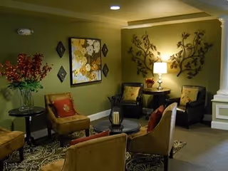 A cozy seating area in a senior living facility with four upholstered chairs arranged around a small round table. The walls are painted green and decorated with floral artwork and metal wall art. A table lamp provides warm lighting, and a vase with red flowers sits on a side table.
