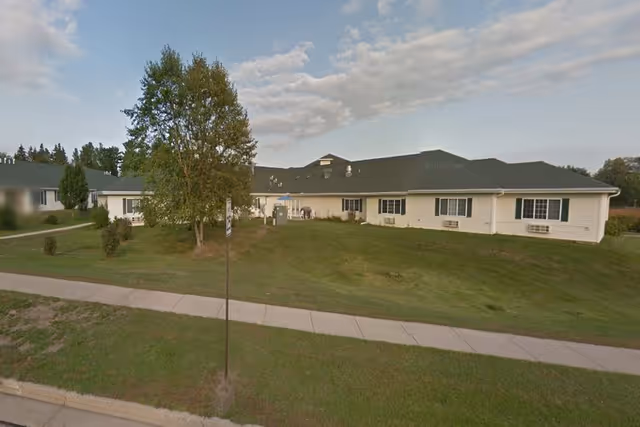 Single-story senior living facility with white siding, green lawn, trees, and a sidewalk.