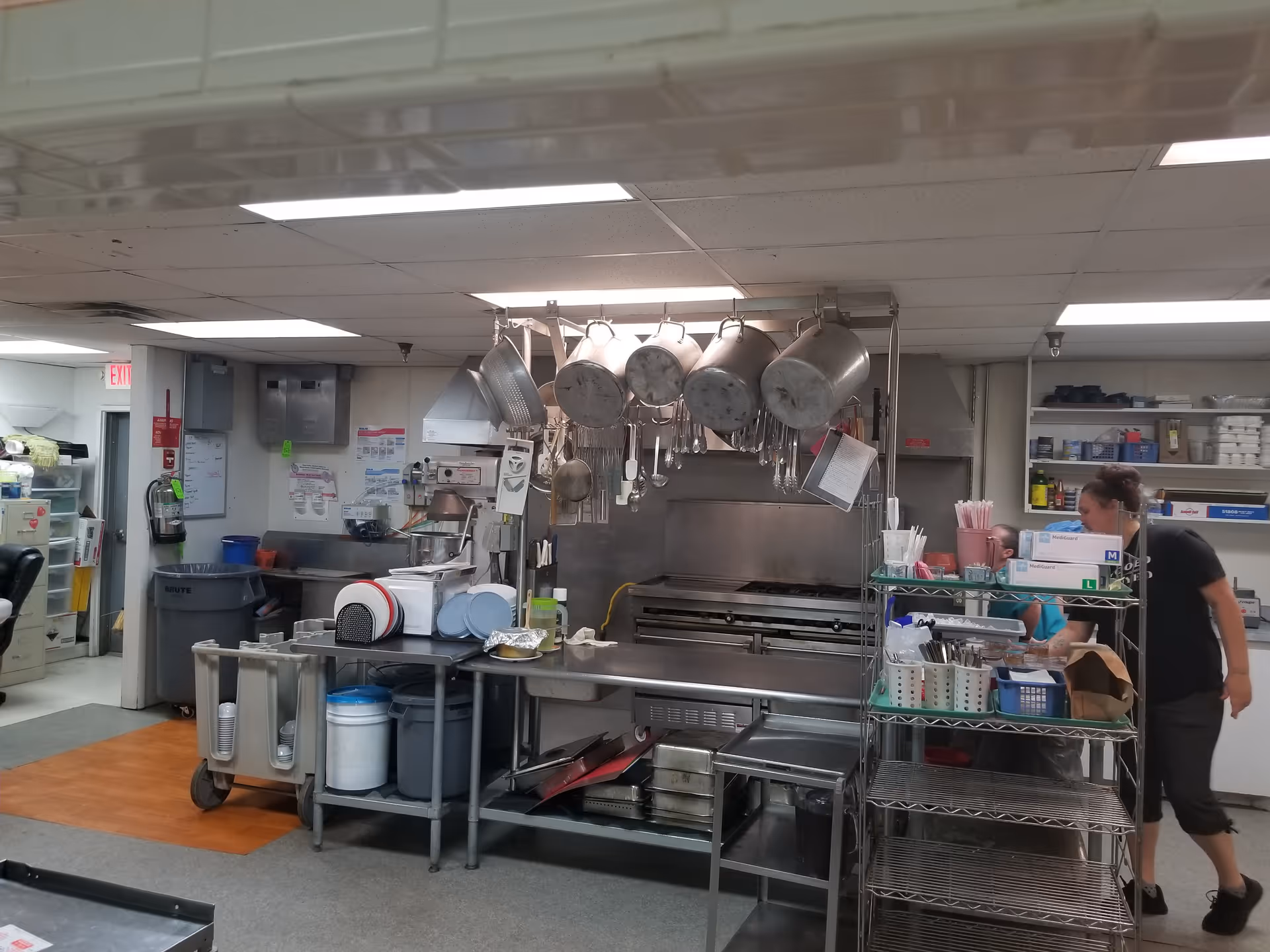A commercial kitchen with pots and pans hanging above a large stove and oven. There are shelves with various kitchen supplies and utensils, and two people are visible working in the background. The kitchen has fluorescent lighting and a mix of gray and wood flooring.