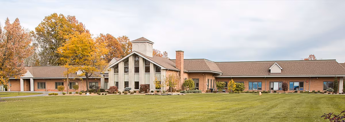 Exterior view of Briarfield Manor, a single-story brick building with large windows and a central tower-like structure, surrounded by a well-maintained lawn and trees with autumn foliage under a cloudy sky.