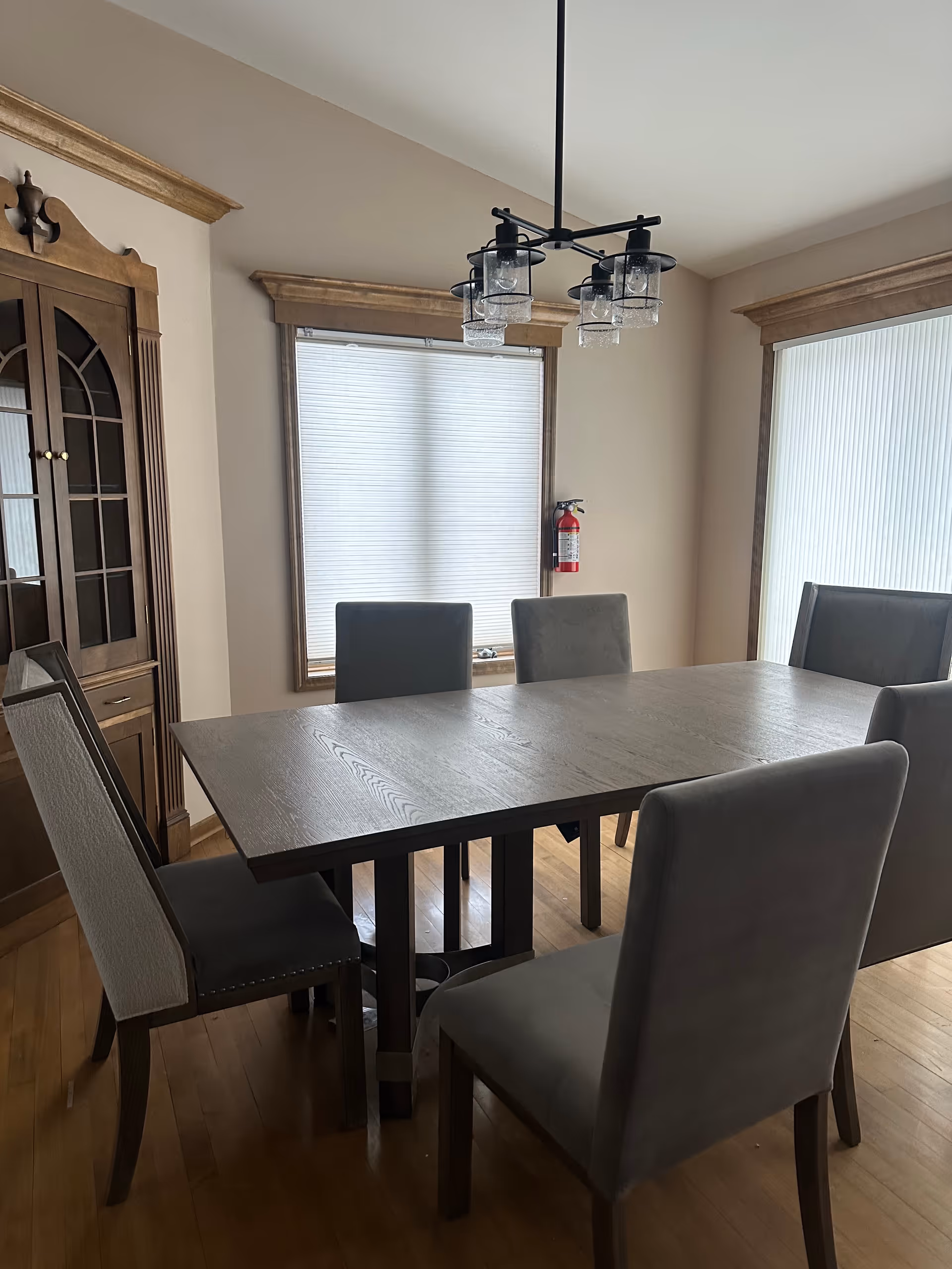 Dining room featuring a large wooden table, upholstered chairs, cabinet, chandelier, and window blinds.