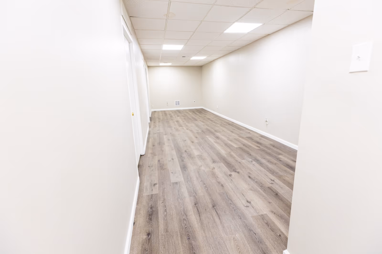 Bright empty hallway with white walls, wood-like flooring, and ceiling lights.
