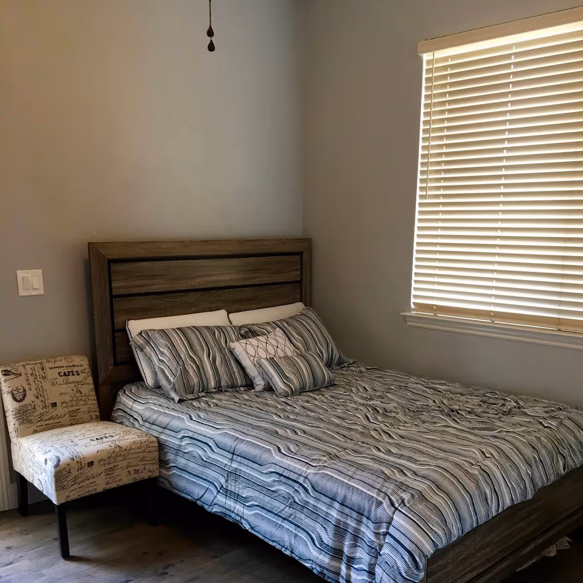 A bedroom with a wooden bed frame and a bed made with striped bedding in shades of gray and white. There are multiple pillows on the bed. Next to the bed is an upholstered chair with a script pattern. A window with closed blinds is on the right wall, and a light switch is visible on the left wall.