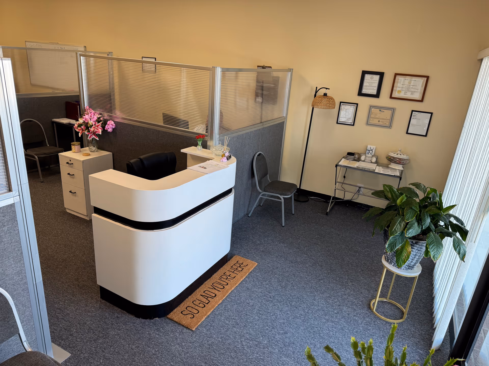 Reception area with a curved white desk, glass dividers, framed certificates, and potted plants.