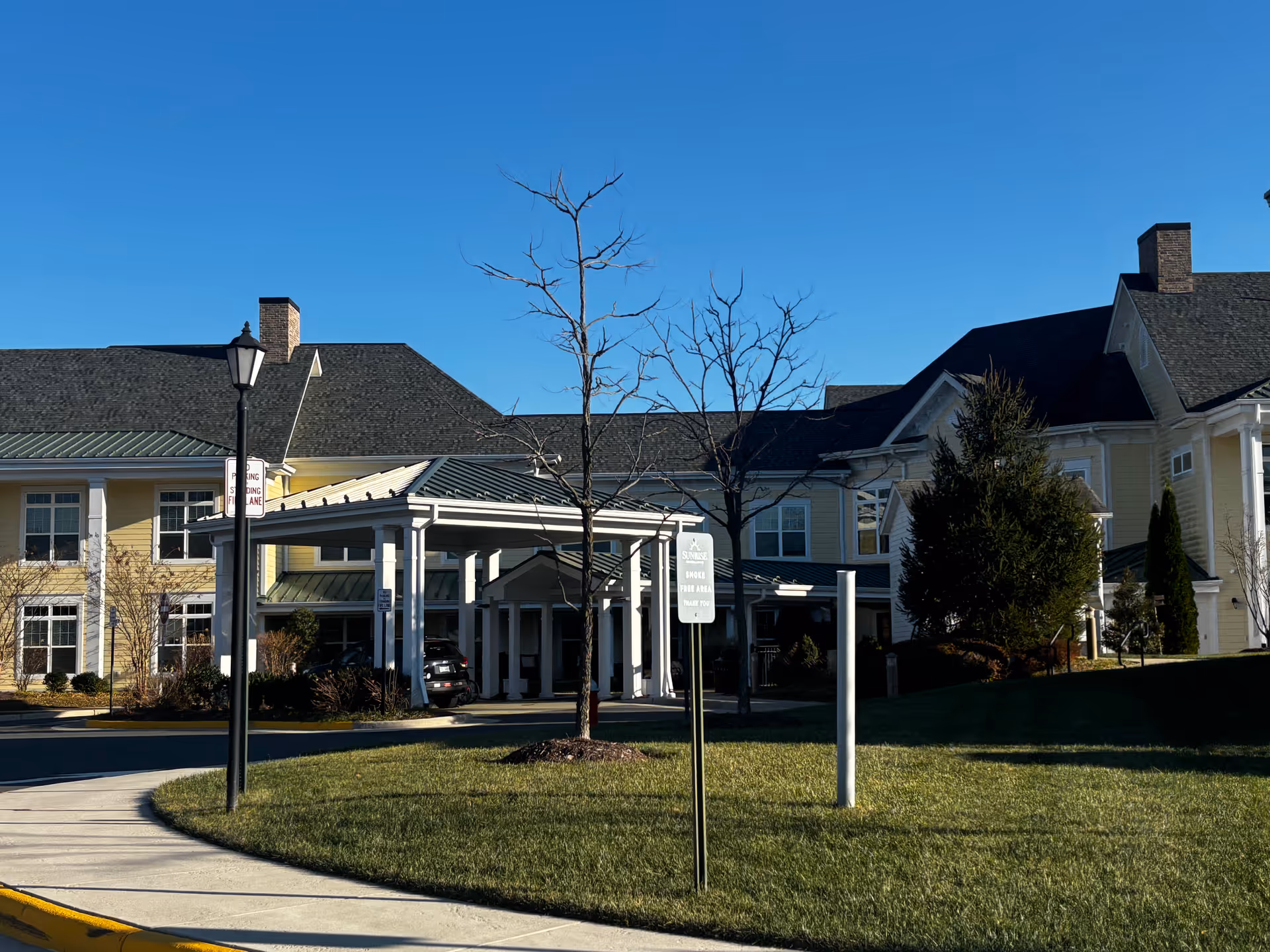 Exterior view of a senior living facility building with a covered entrance, a lamp post, leafless trees, and a green lawn under a clear blue sky.