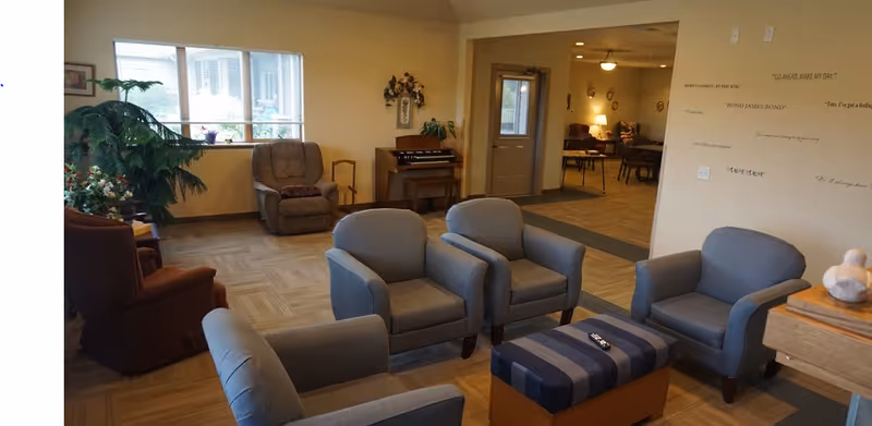 A cozy living room area in an assisted living facility featuring four gray armchairs arranged around a striped ottoman. In the background, there is a brown recliner, a small organ, and a large window letting in natural light. The walls are decorated with inspirational quotes, and there are plants adding greenery to the space. The room opens into another area with more seating and warm lighting.