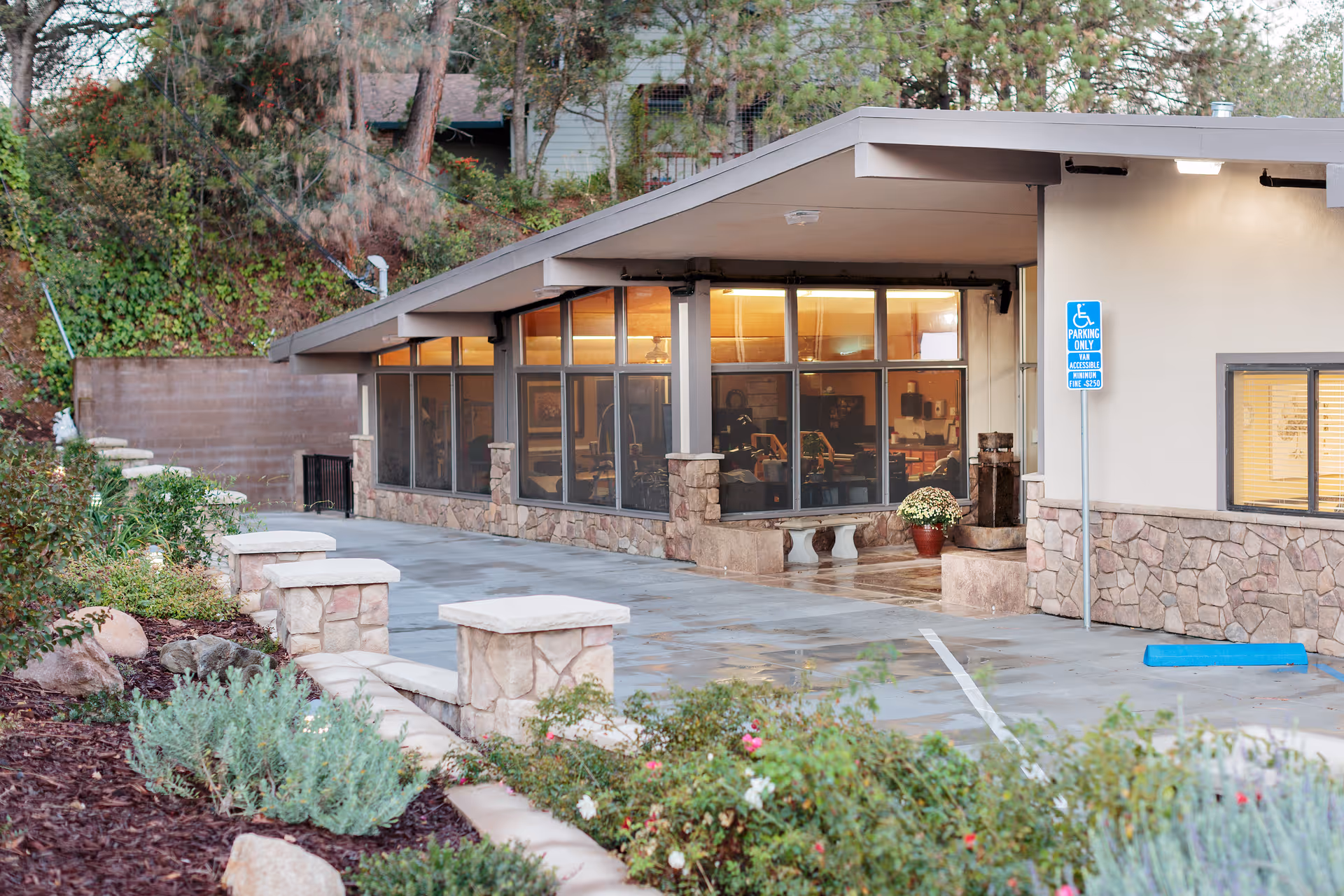 Exterior view of a building with large windows and stone accents, surrounded by landscaped plants and a paved walkway. There is a handicapped parking sign and a blue parking space visible near the entrance.