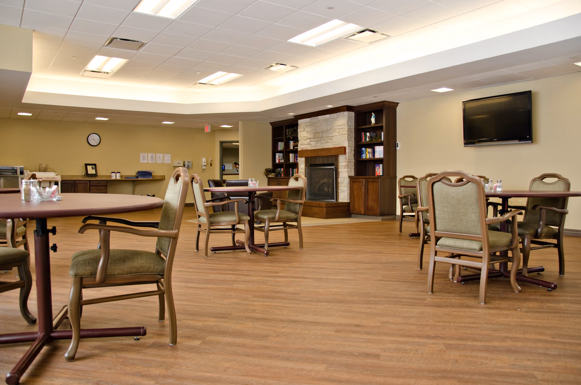 A spacious dining room with several round tables and cushioned chairs arranged on a wooden floor. There is a stone fireplace with built-in wooden shelves filled with books and decorative items. A flat-screen TV is mounted on the wall to the right. The room has a drop ceiling with recessed lighting and a clock on the far wall above a counter area.