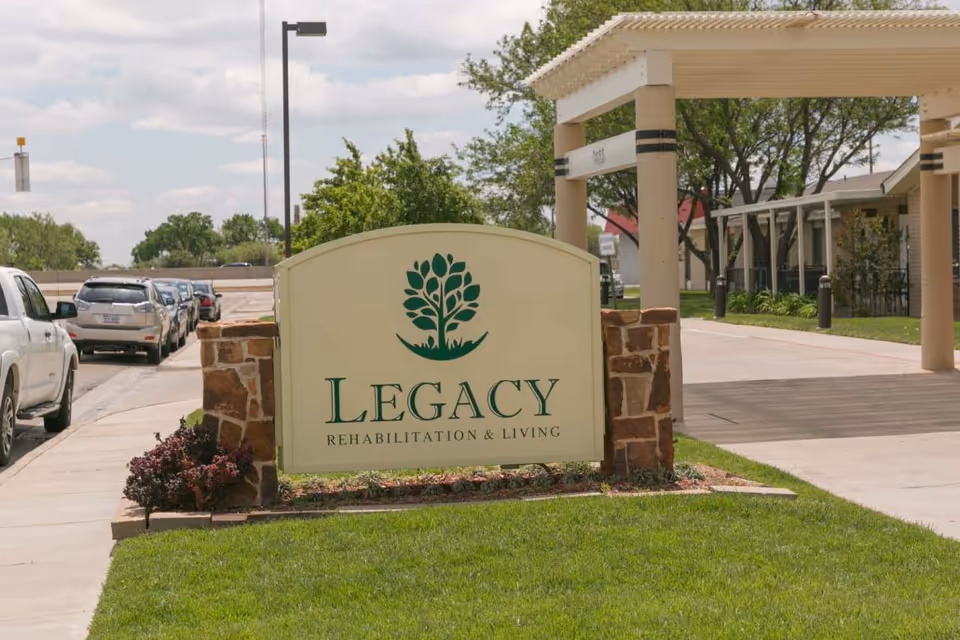 Legacy Rehabilitation and Living entrance sign with stone posts, green lawn, and parked cars.