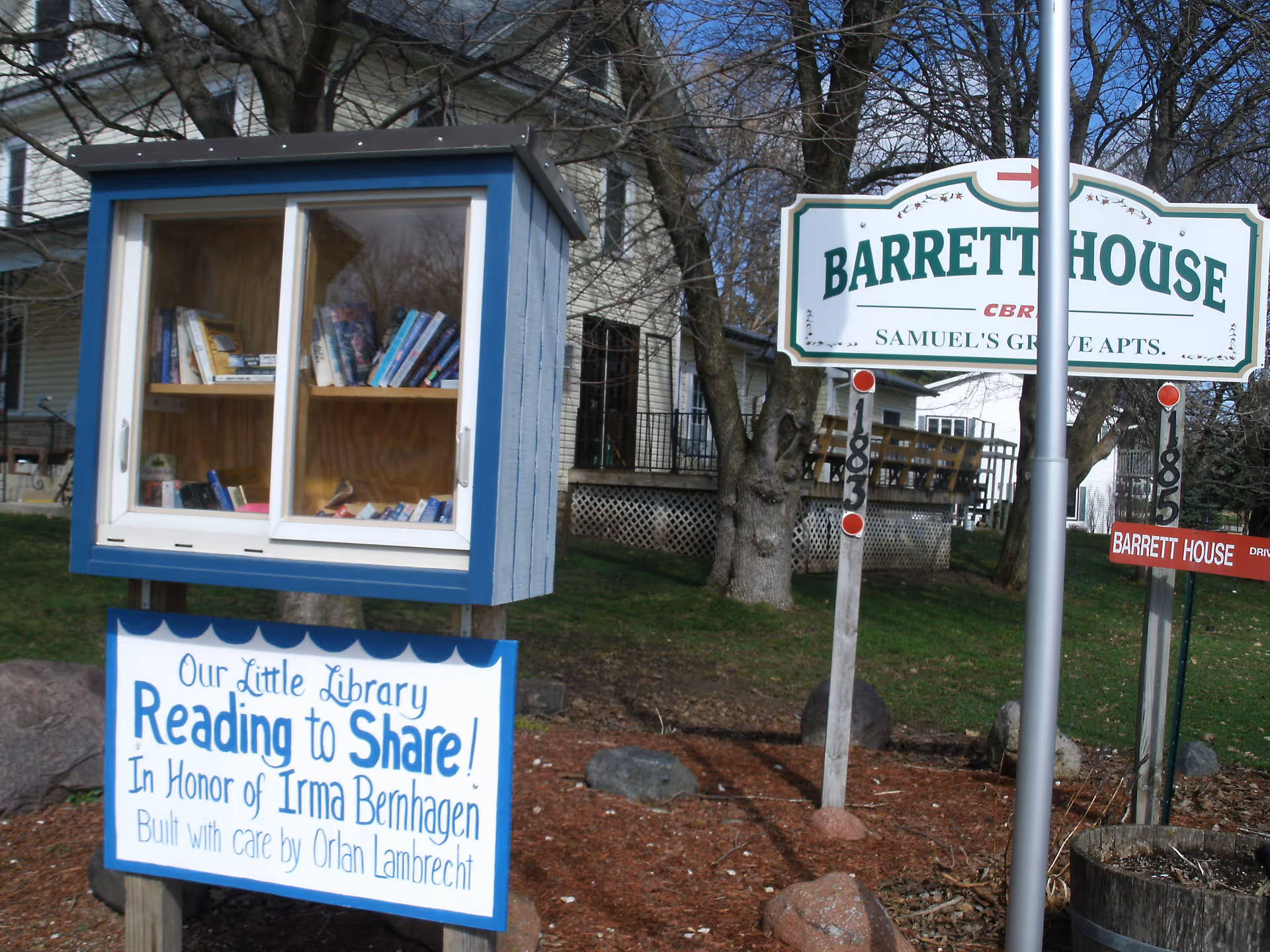 A small outdoor little free library box filled with books next to a sign for Barrett House, Samuel's Grove Apartments. The little library sign reads 'Our Little Library Reading to Share! In Honor of Irma Bernhagen Built with care by Orlan Lambrecht'. Trees and a building are visible in the background.