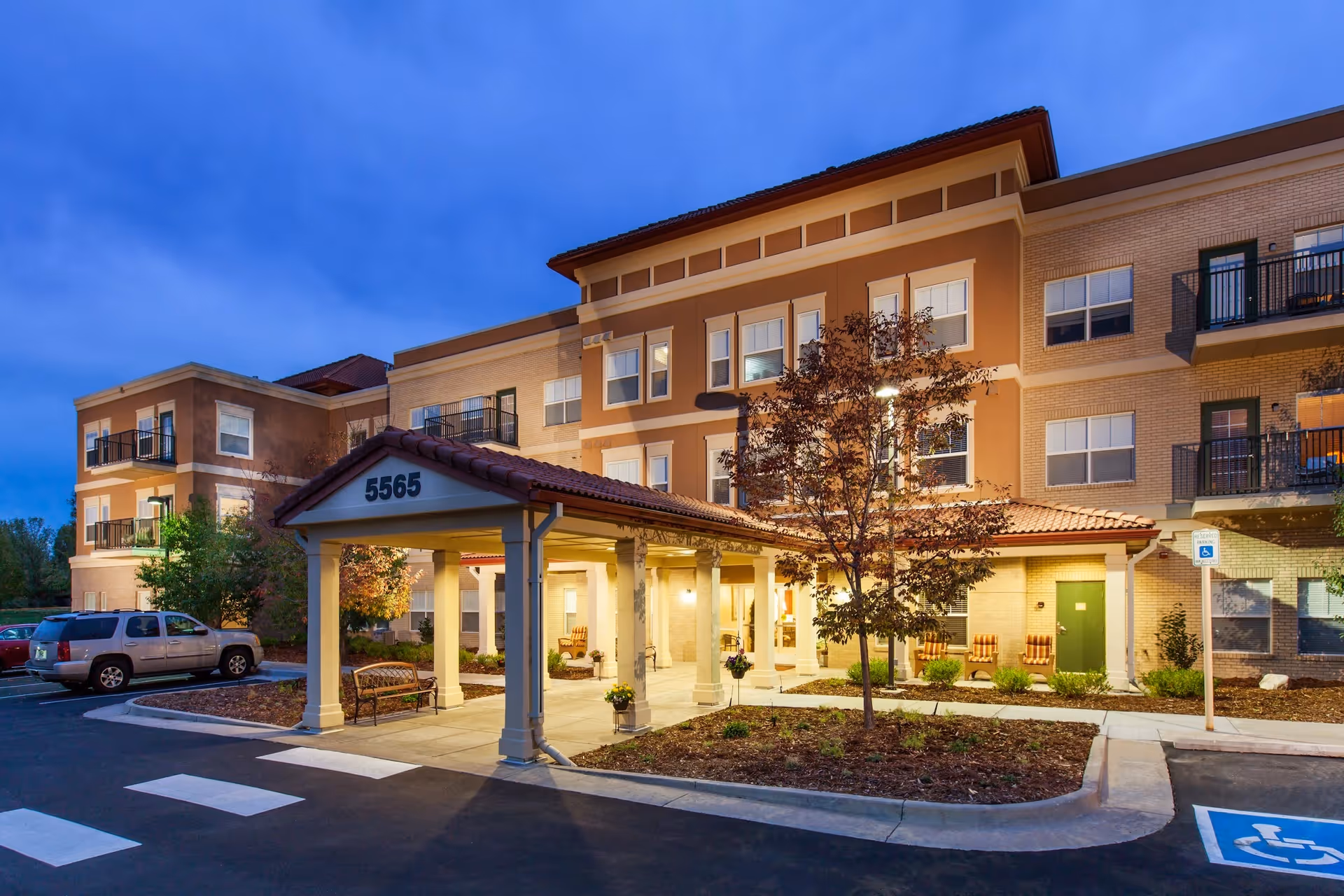 Exterior view of a multi-story senior living facility building at dusk with a covered entrance marked 5565, surrounded by landscaped areas and a parking lot with handicap parking spaces.
