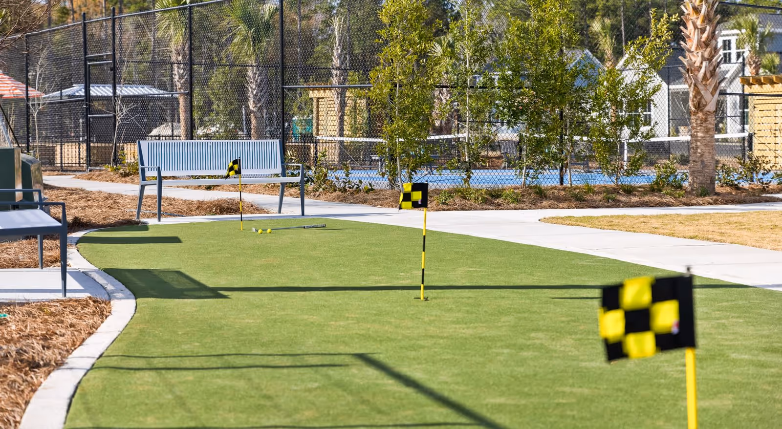 Outdoor putting green area with three yellow and black checkered flags, a golf putter, and three golf balls. There are benches along the paved walkway, trees, and a chain-link fence in the background.