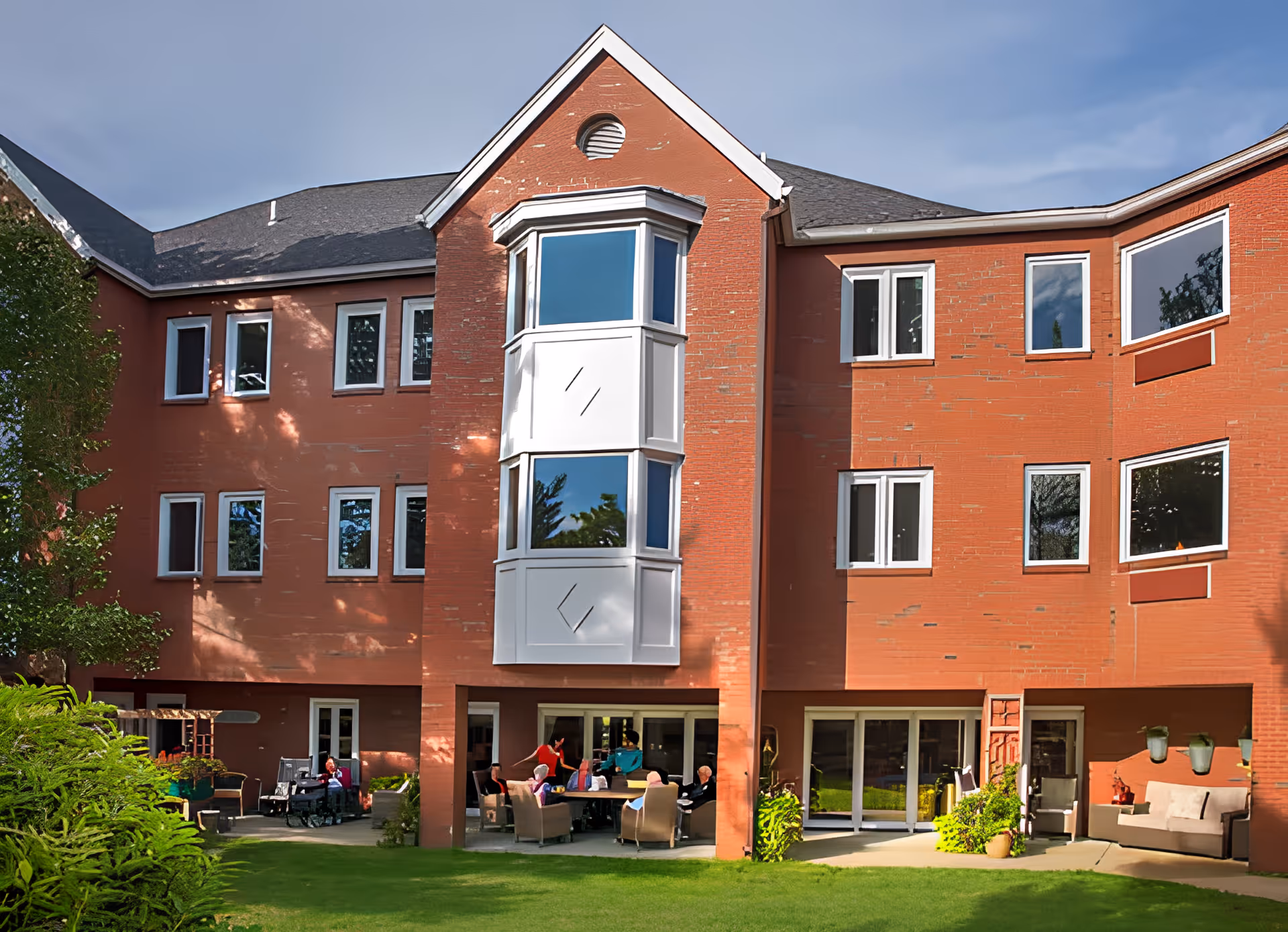 Three-story red-brick senior living building with a central bay window and ground-level patio where residents sit on outdoor furniture facing a lawn.