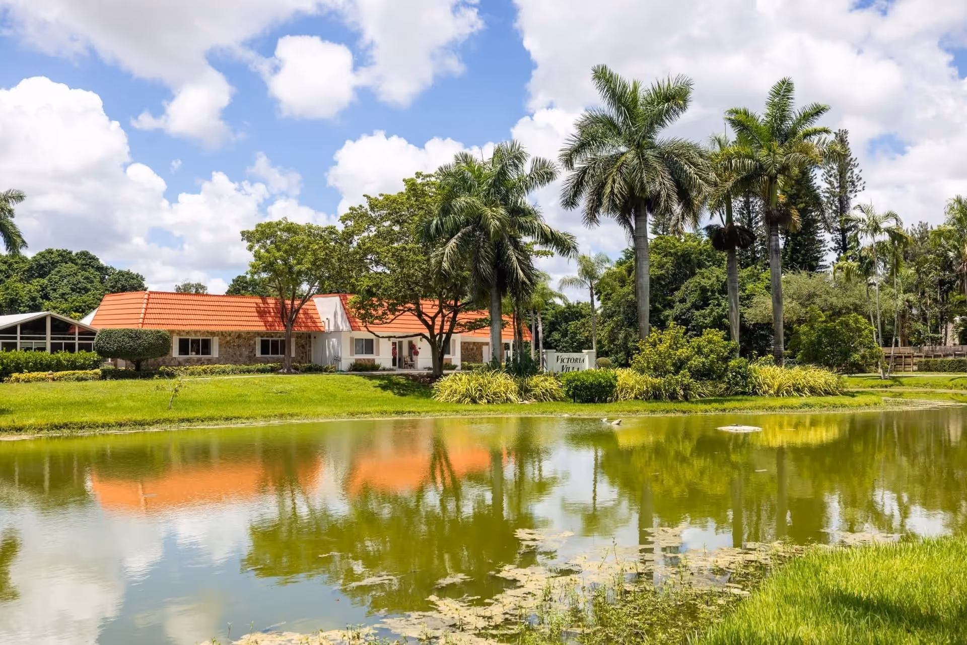 Victoria Villa Assisted Living building with a red roof, palm trees, and a reflecting pond.