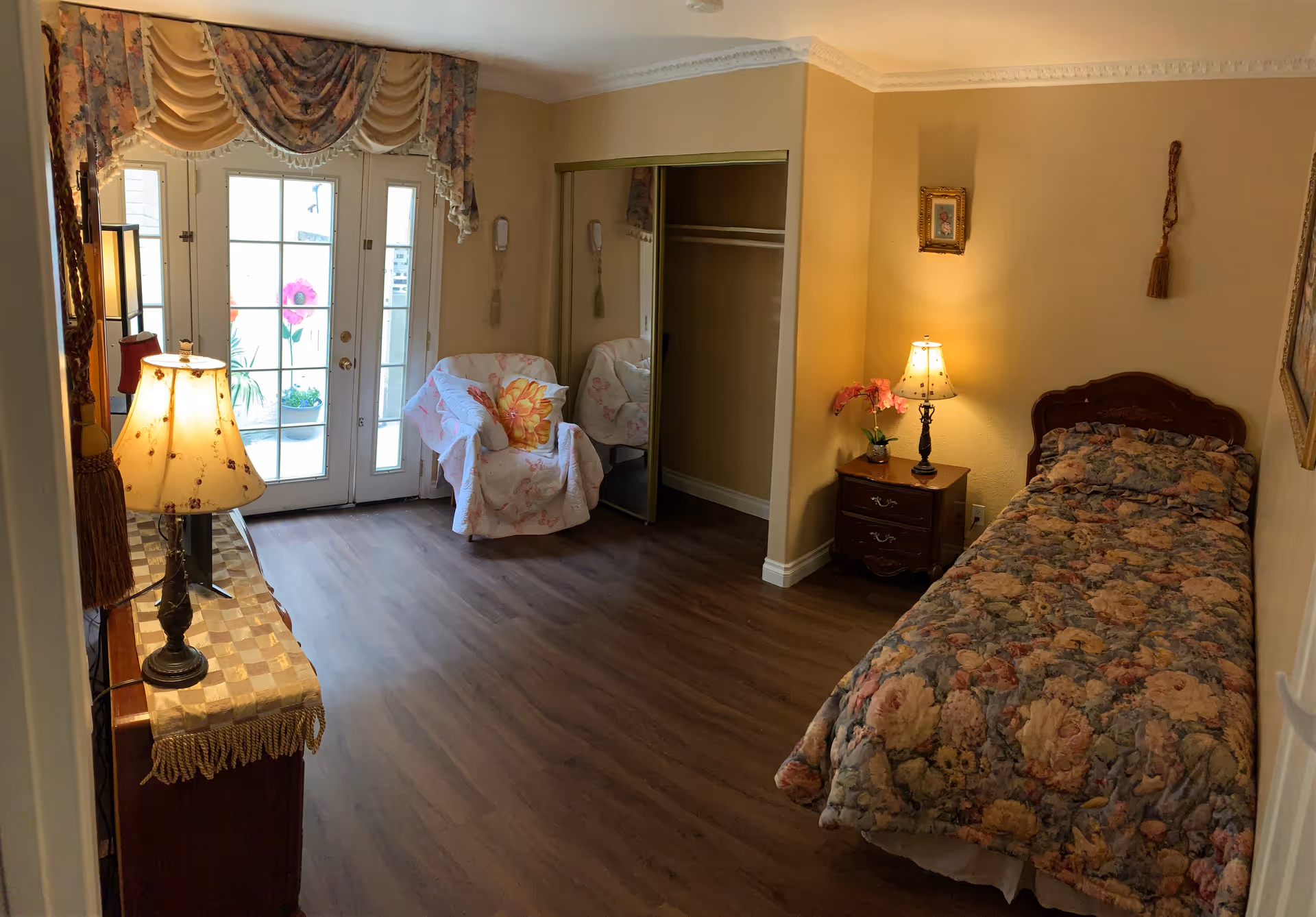 Small furnished bedroom with floral bedspread, lamps, nightstand, and a seating area near French doors.