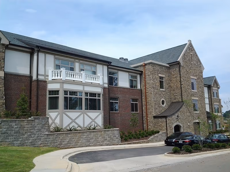 Front exterior of a stone-and-brick multi-story senior living building with a driveway and parked cars.