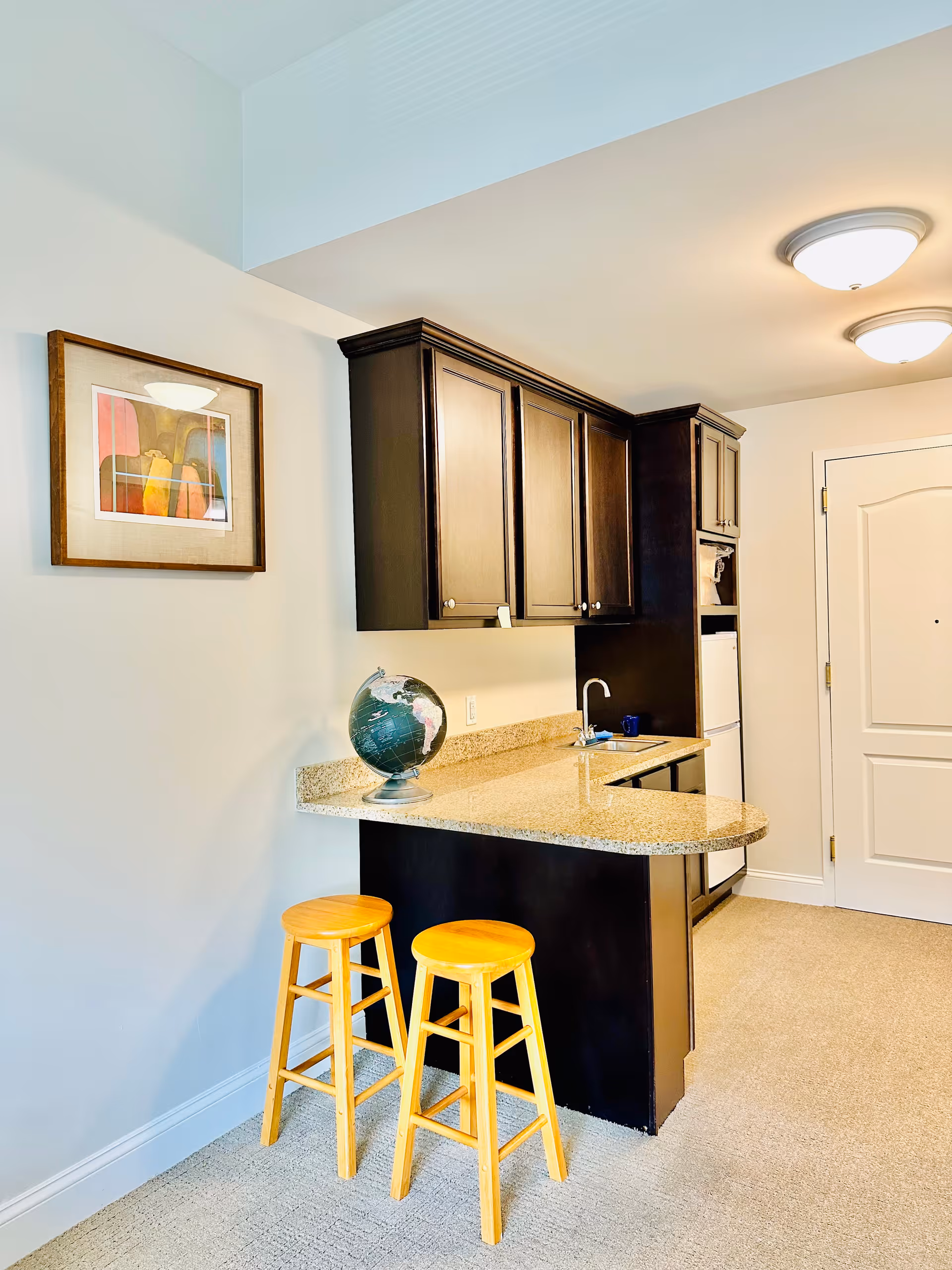 Bright, modern kitchenette with dark cabinets, granite counter, stools, globe, and sink.