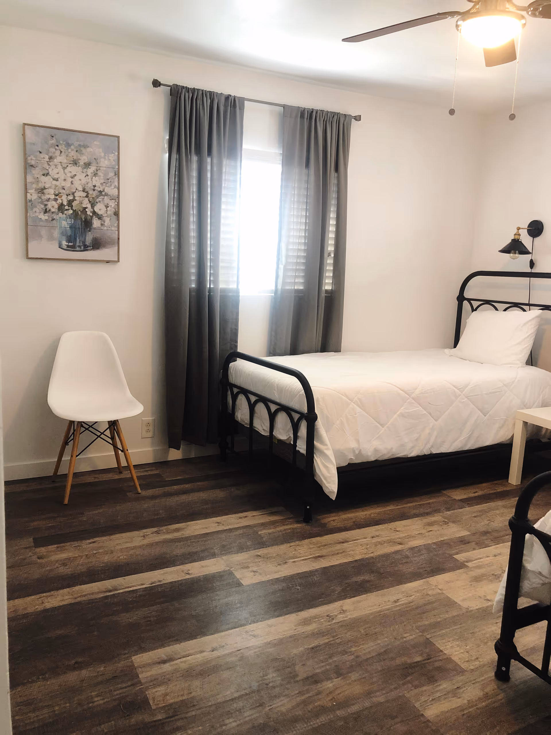 Bright bedroom with a single black metal-framed bed, white bedding, a white chair, grey curtains over a window, and wood-look flooring.