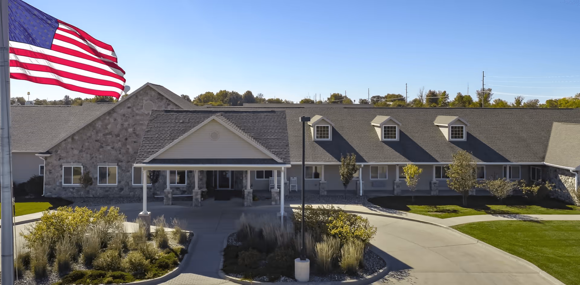 Exterior view of Homestead Assisted Living & Memory Care of Osceola building with a large American flag on a flagpole in the foreground, a covered entrance, stone and siding facade, and landscaped greenery under a clear blue sky.