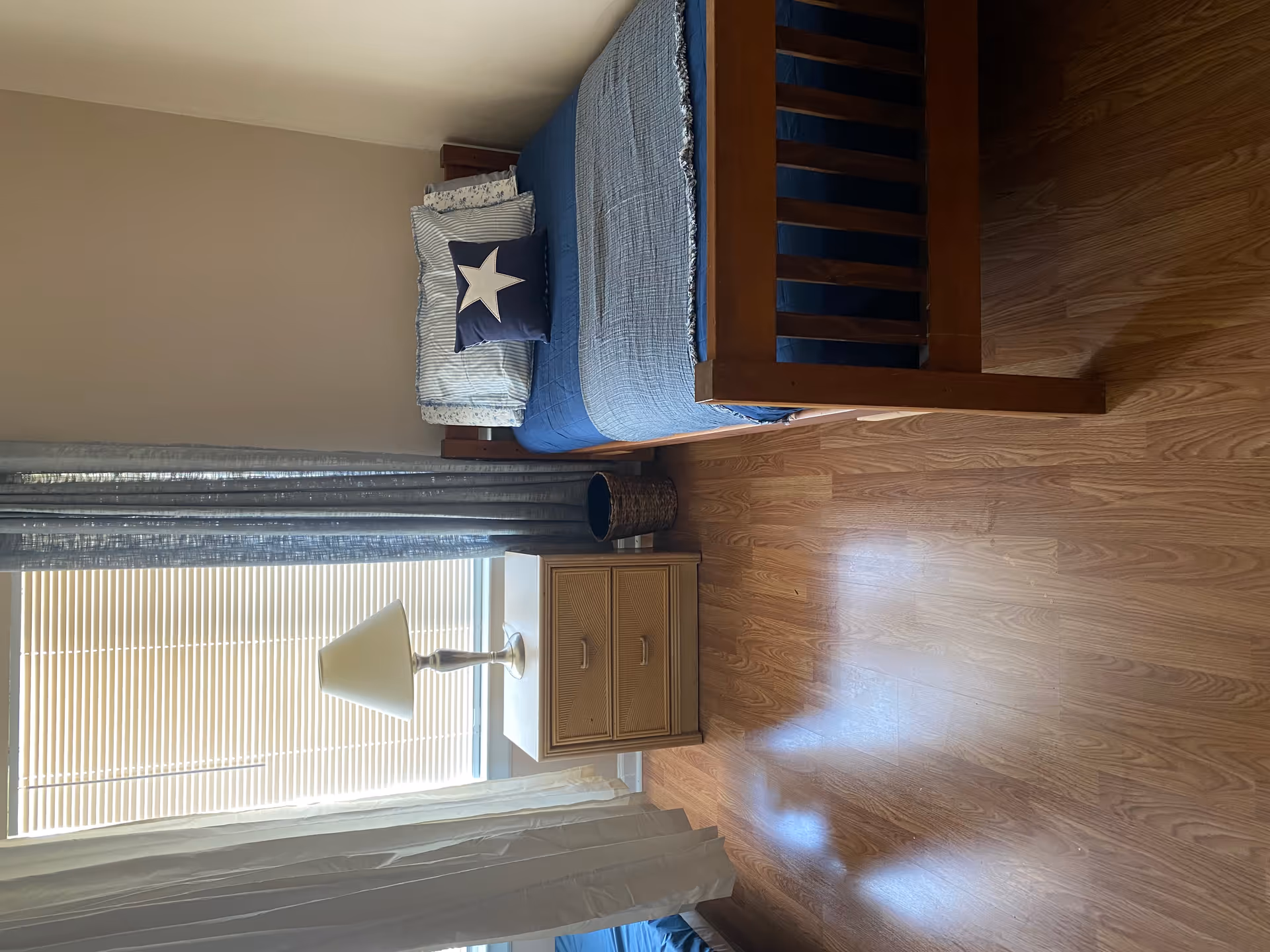 Single bedroom with wooden bed frame, blue bedding, nightstand, lamp, and window with blinds.