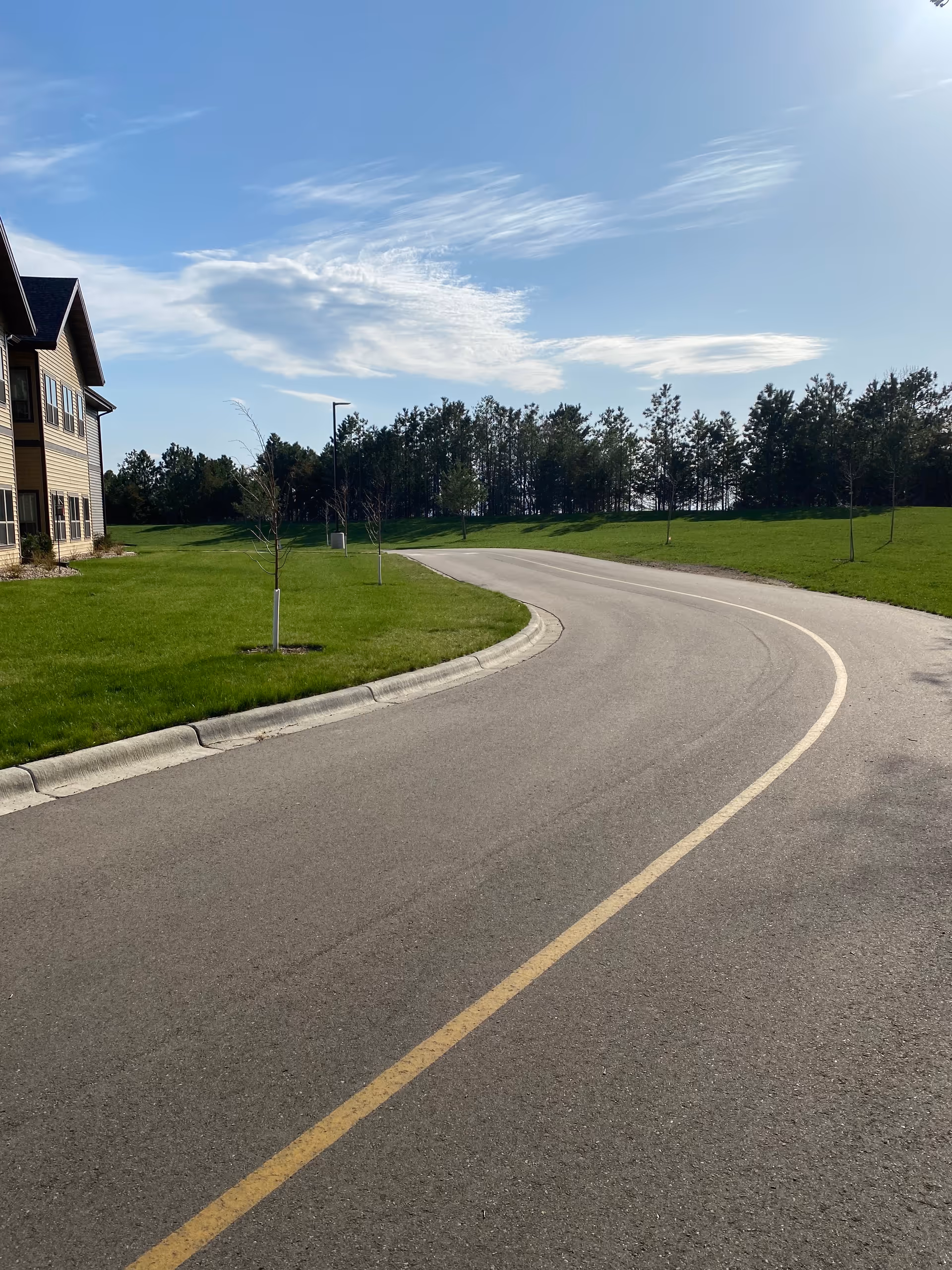 Curved road curves past a beige residential building with green lawns and trees under blue sky.