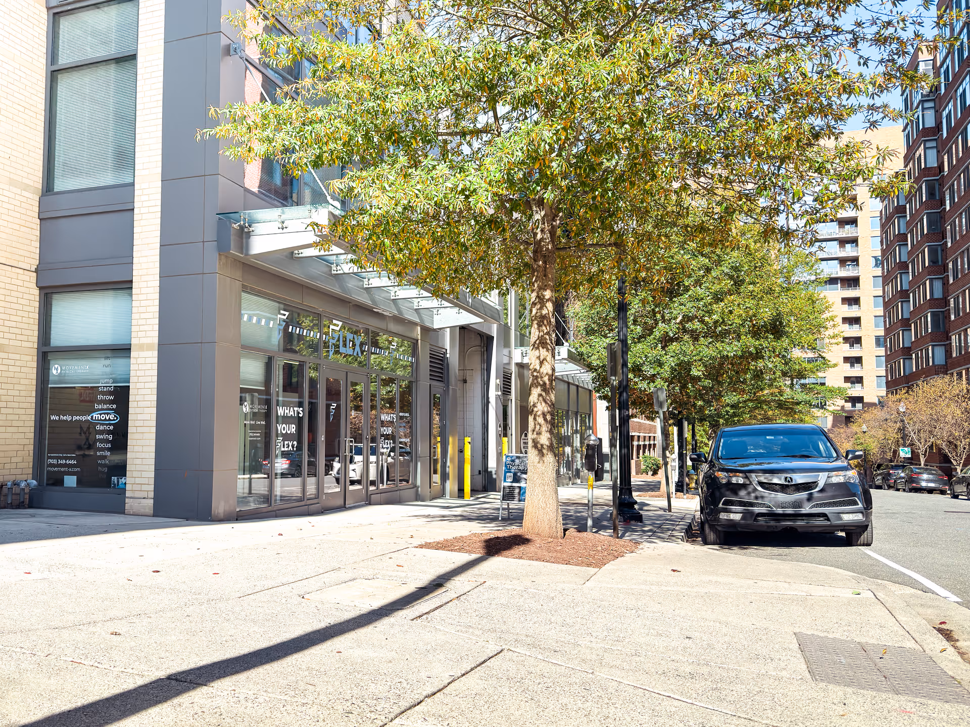 Street view of a modern building entrance with glass doors, trees, and a parked car.