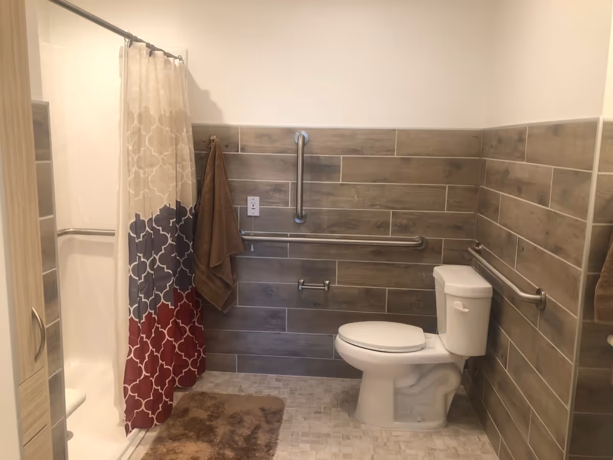 A bathroom with a white toilet, beige and brown tiled walls, a shower with a patterned curtain in shades of beige, gray, and red, brown towels hanging on hooks, and safety grab bars installed on the walls around the toilet and shower area. There is a brown bath mat on the tiled floor.