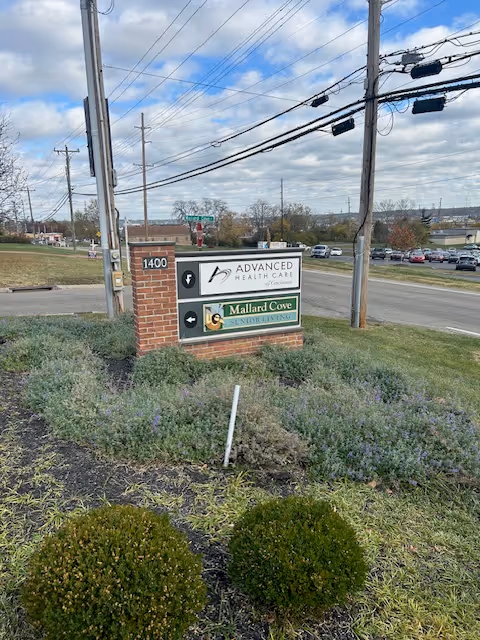 Brick sign displays multiple facility names near a road with landscaping.